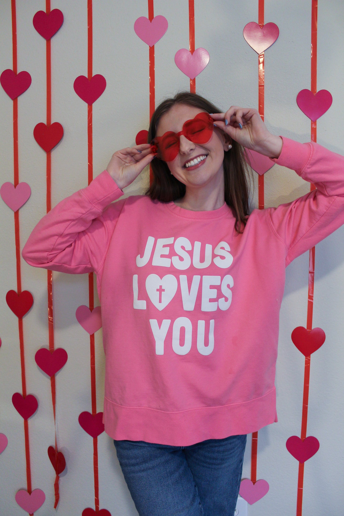 Person wearing a pink sweatshirt with 'Jesus Loves You' text, posing with heart-shaped sunglasses against a heart-patterned background.