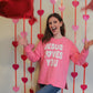 Person wearing a pink sweatshirt with 'Jesus Loves You' text, standing in front of heart decorations.
