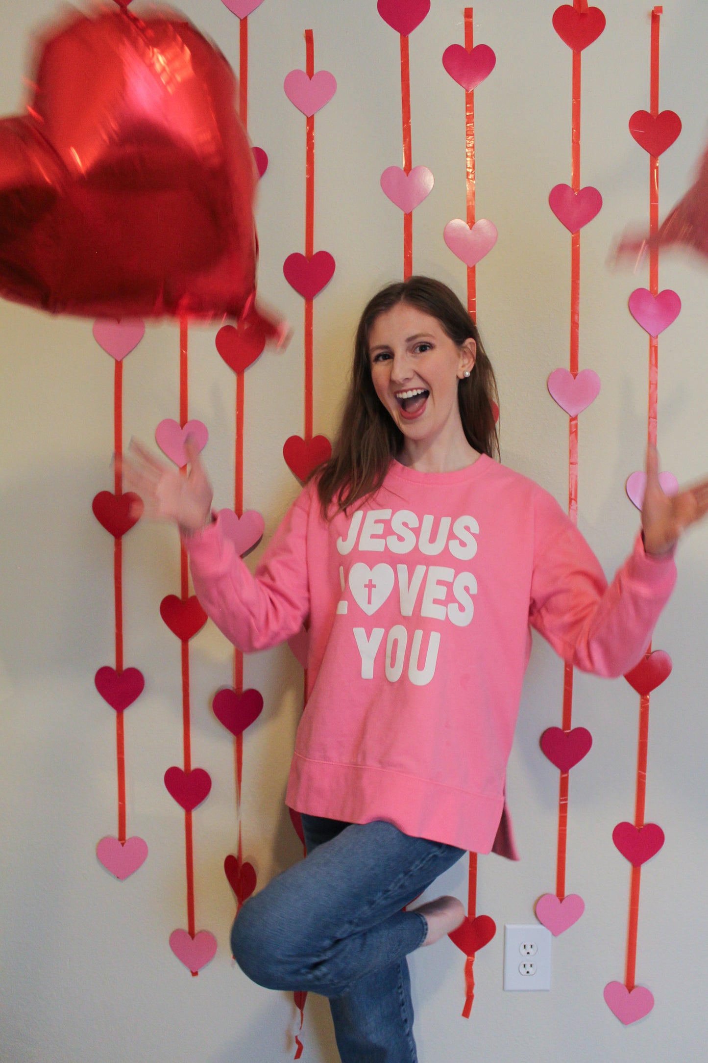 Person wearing a pink sweatshirt with 'Jesus Loves You' text, standing in front of heart decorations.